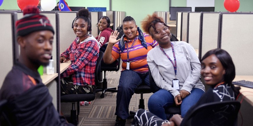 A group of contact center agents lean out of their cubicles and smile in a large office.