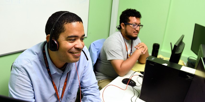 Two men sit at their desks with headsets on, smiling at their computer screens.