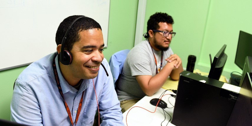 Two men sit at desks in front of monitors wearing headsets.